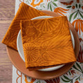An orange patterned fabric napkin with a yellow-orange floral and leaf design is folded over a white plate on a brown ceramic plate, set on a matching patterned table runner.