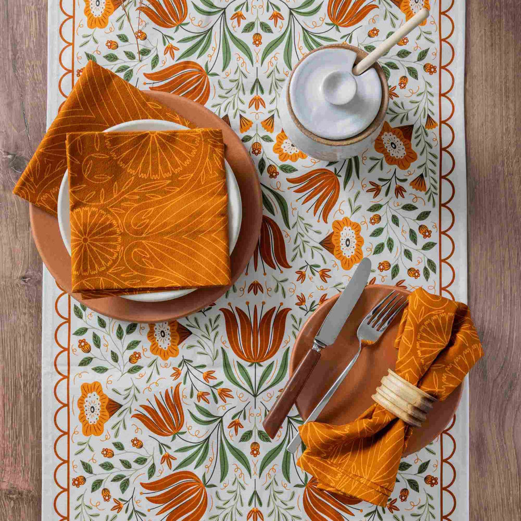 An overhead flat-lay of a place setting on a patterned table runner. The setting includes terracotta-colored plates, a white ceramic lidded container with a wooden spoon, silver-toned cutlery, and two folded orange napkins with a matching orange floral print.