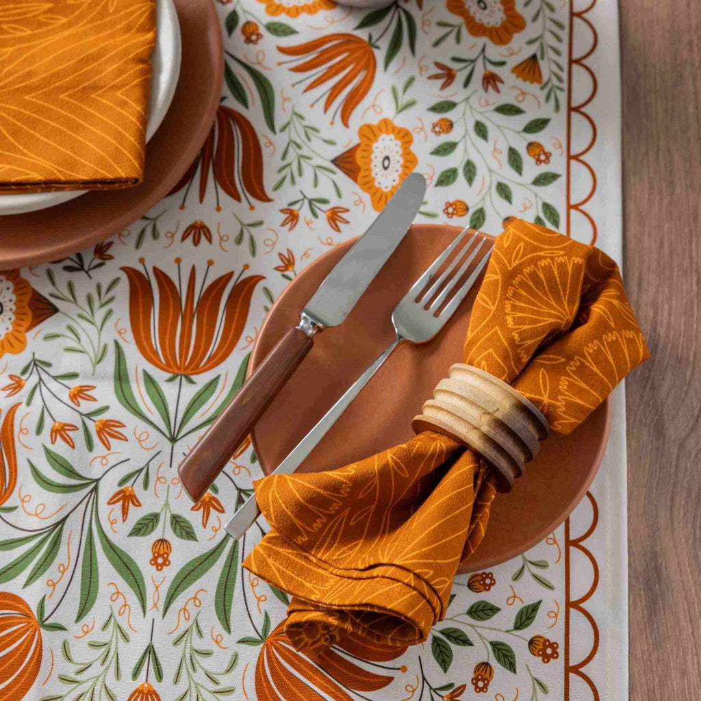 An autumn-themed table setting featuring a terracotta-colored plate, silver cutlery, a folded orange napkin with a white leaf pattern, and a matching napkin under a wooden napkin ring, all arranged on a white tablecloth with a bold orange and green floral tulip pattern.