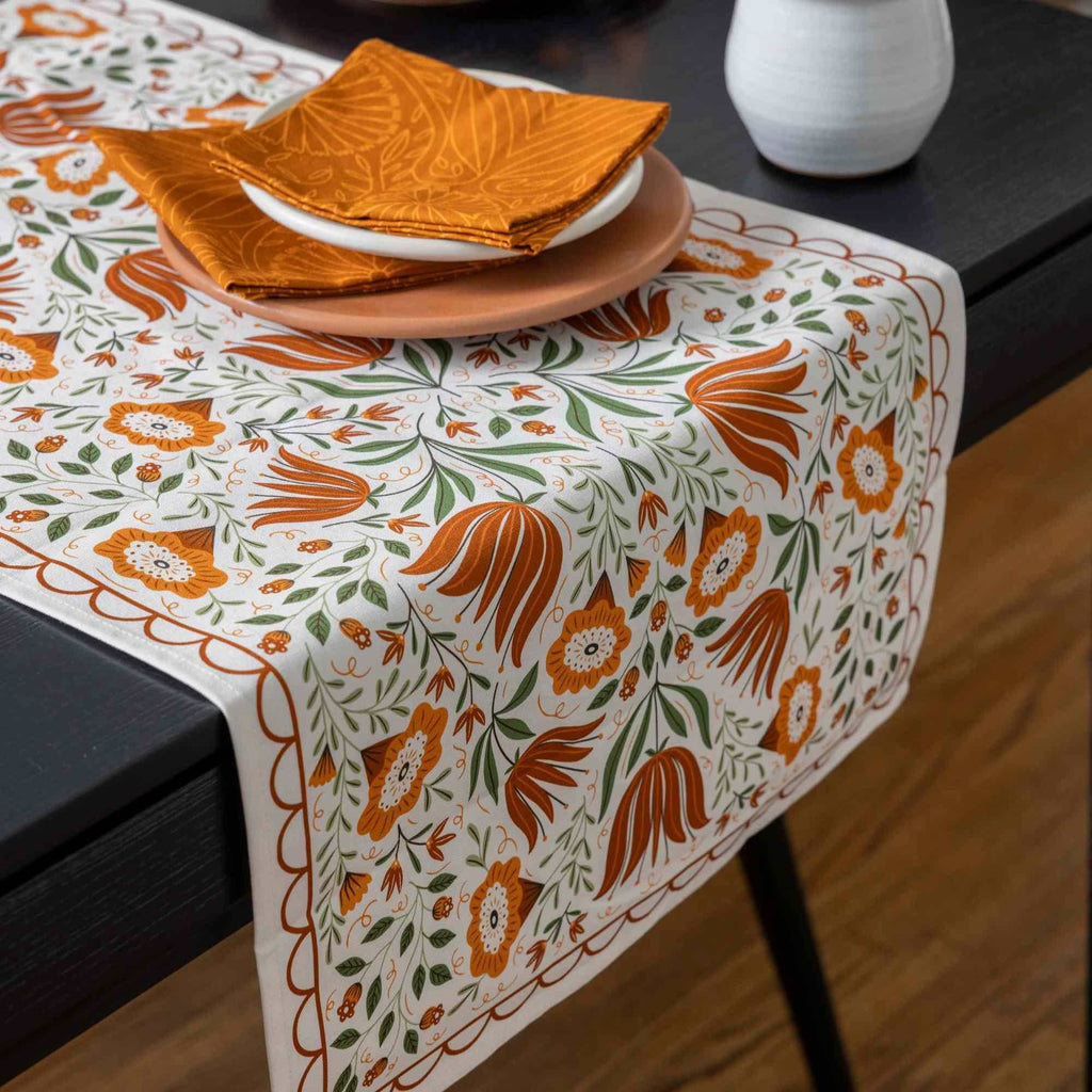 A rectangular table runner with an orange, green, and white floral pattern draped over a dark table, styled with plates and folded napkins.
