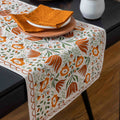 A rectangular table runner with an orange, green, and white floral pattern draped over a dark table, styled with plates and folded napkins.