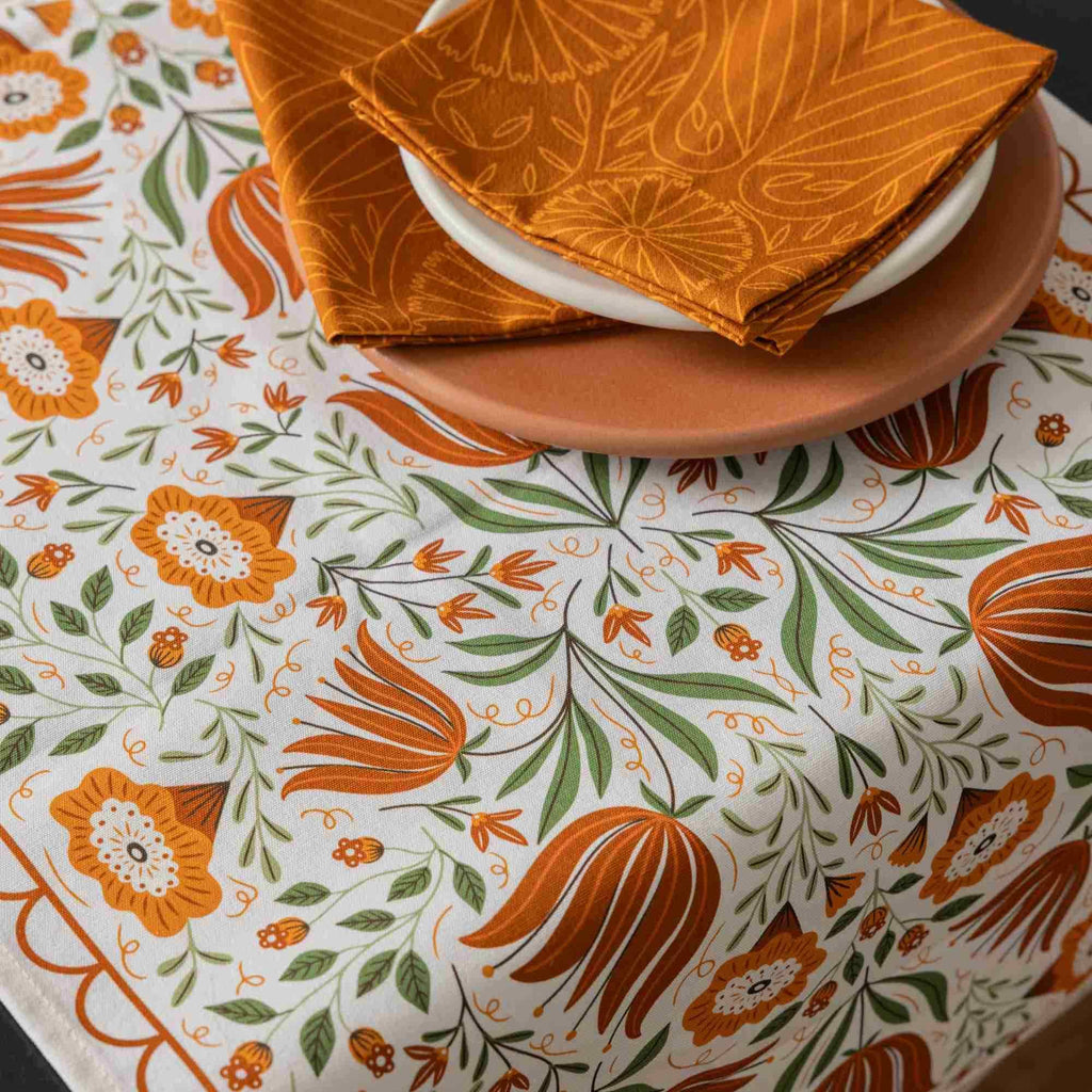 A close-up of a white tablecloth with a colorful botanical pattern in orange and green, styled with a terracotta plate and folded orange napkins on a white table.