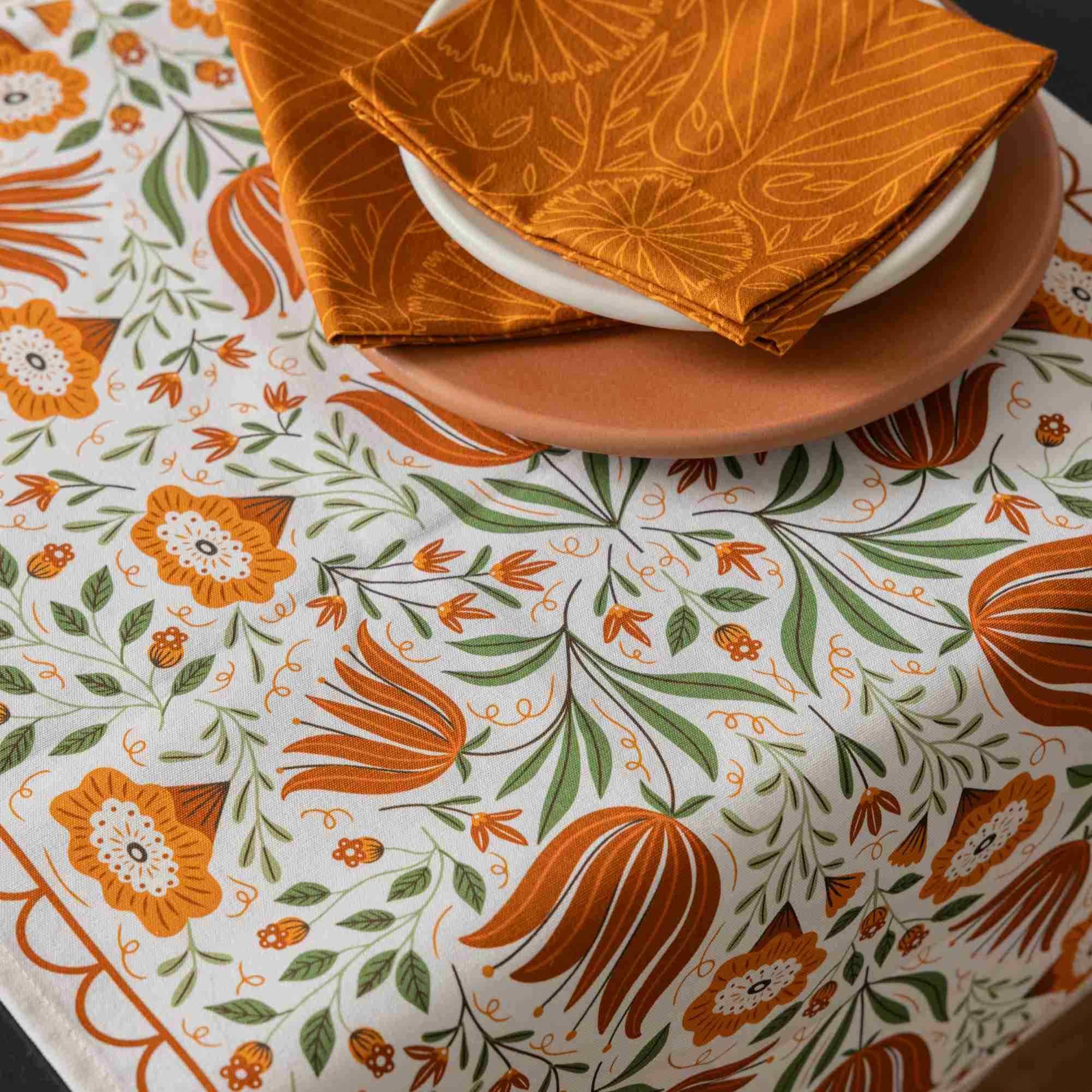 A close-up of a white tablecloth with a colorful botanical pattern in orange and green, styled with a terracotta plate and folded orange napkins on a white table.