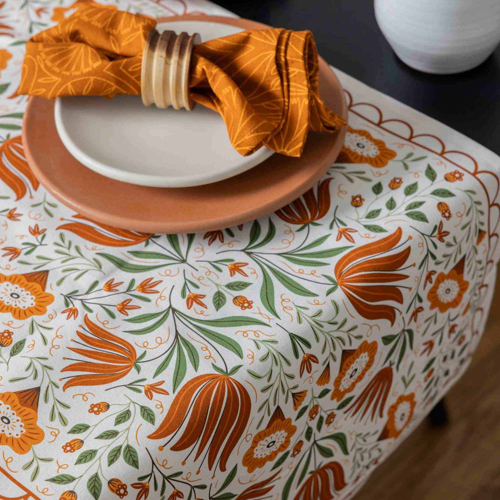 A close-up of a table setting featuring a white tablecloth with a vibrant orange and green botanical pattern, styled with a terracotta plate, a white plate, and an orange napkin tied with a wooden ring.