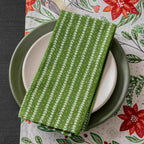 A green cotton patterned napkin with a white floral pattern is draped over a stack of plates, set on a festive Christmas table runner with poinsettias and birds.