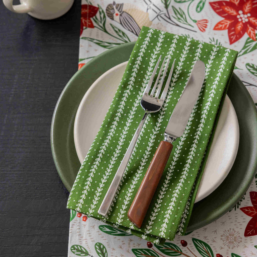 A styled tabletop setting featuring a green and white patterned napkin with silver cutlery, layered over white and green plates, on a festive holiday table runner with a bird and floral print.