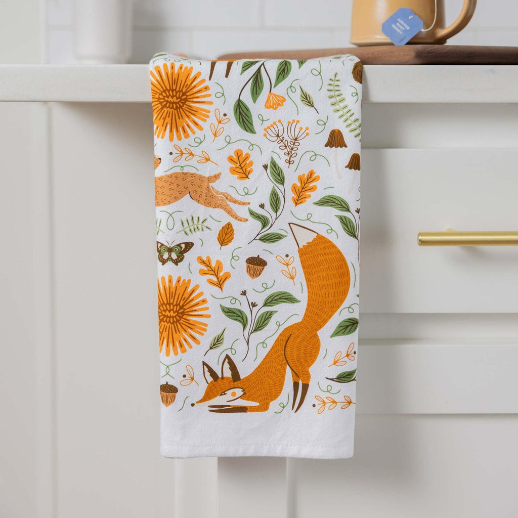 A white kitchen towel with a colorful autumn-themed print of a fox, sunflowers, leaves, acorns, and a butterfly, hanging from a white cabinet handle.