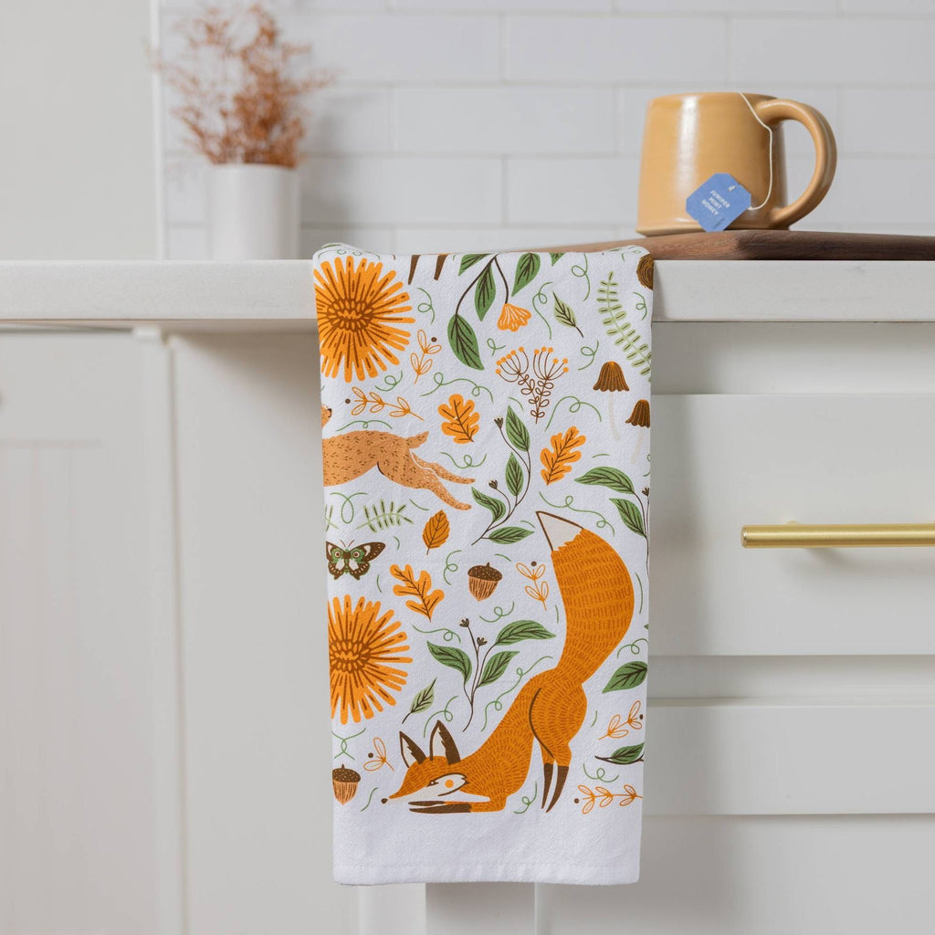 A white kitchen towel with a colorful autumn forest pattern featuring a fox, sunflowers, acorns, and leaves, hanging from a white cabinet handle in a kitchen setting.