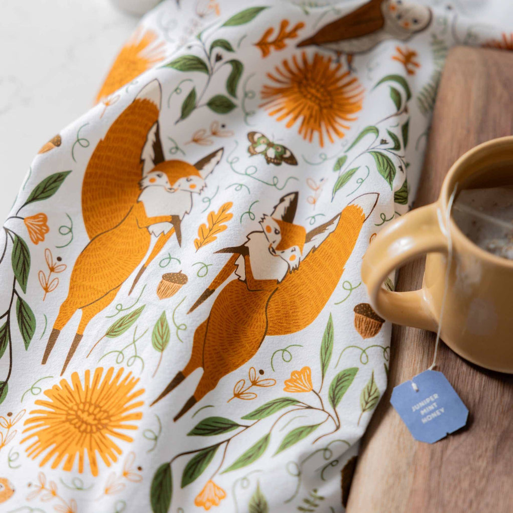 A white tea towel with a printed pattern of orange foxes, yellow flowers, and green leaves, styled next to a ceramic mug on a wooden surface.