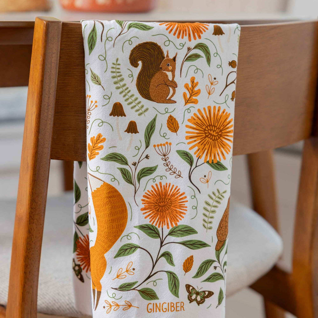 A white kitchen tea towel with an autumn-themed print of a squirrel, orange flowers, and green leaves, draped over the back of a wooden chair. The word 'GINGIBER' is printed at the bottom.
