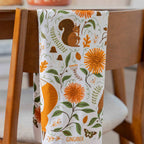 A white kitchen tea towel with an autumn-themed print of a squirrel, orange flowers, and green leaves, draped over the back of a wooden chair. The word 'GINGIBER' is printed at the bottom.