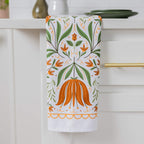 A white kitchen towel with a symmetrical botanical pattern of green leaves and orange flowers, hanging from a gold handle on a white cabinet.