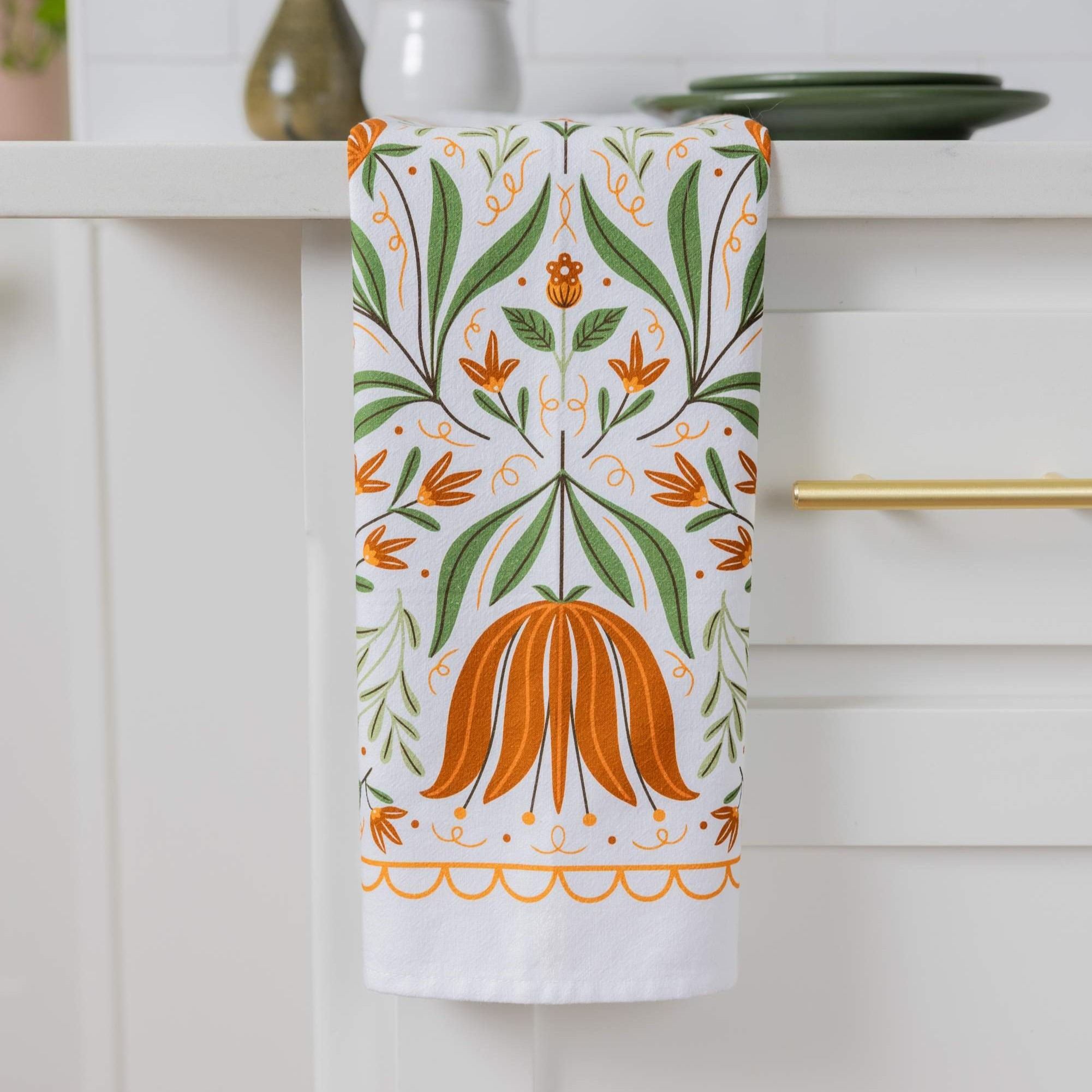 A white kitchen towel with a symmetrical botanical pattern of green leaves and orange flowers, hanging from a gold handle on a white cabinet.