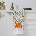 A white kitchen towel with an orange and green botanical pattern hanging from a brass handle on a white kitchen cabinet.