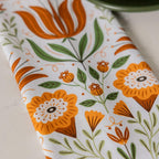 A close-up of a white fabric item, likely a tea towel or napkin, featuring a retro floral pattern in orange, green, and brown.