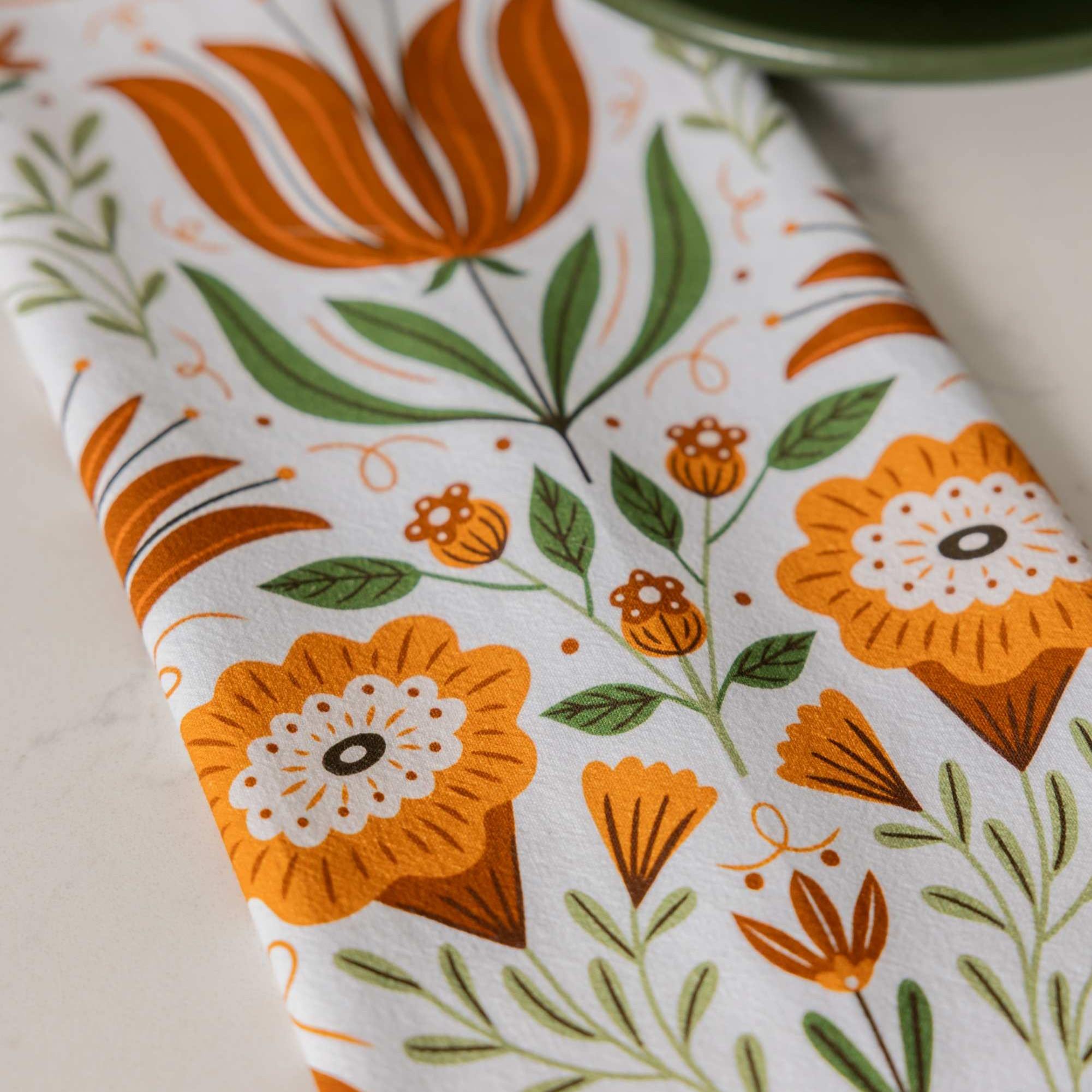 A close-up of a white fabric item, likely a tea towel or napkin, featuring a retro floral pattern in orange, green, and brown.