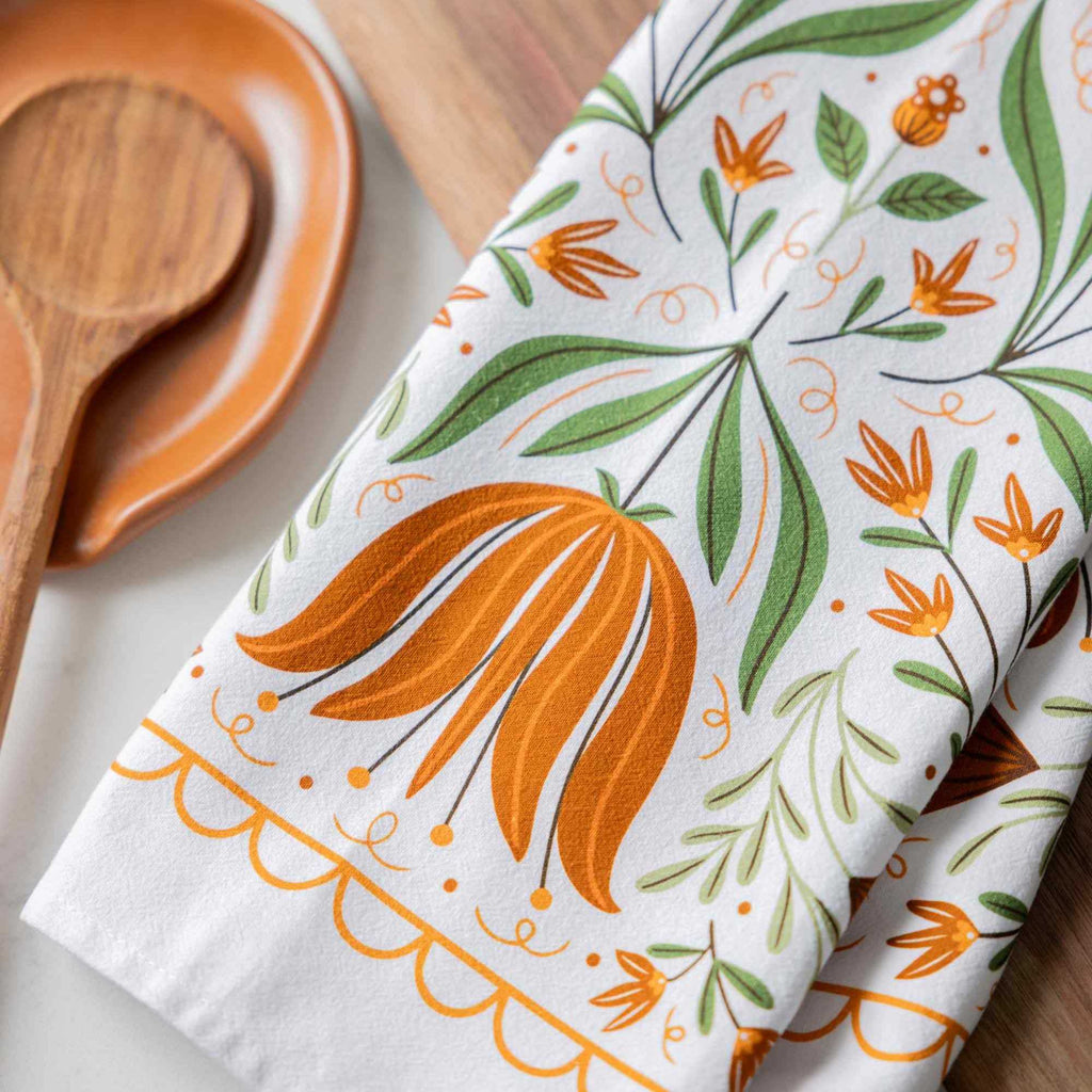 A folded white kitchen towel with a colorful botanical pattern of orange flowers and green leaves, styled on a wooden surface next to a wooden spoon and dish.