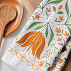 A folded white kitchen towel with a colorful botanical pattern of orange flowers and green leaves, styled on a wooden surface next to a wooden spoon and dish.