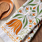 A folded white kitchen towel with a botanical pattern of orange flowers and green leaves, styled on a wooden surface next to a wooden spoon and dish.