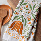 A white kitchen towel with a colorful botanical pattern of orange flowers and green leaves, styled on a wooden cutting board next to a wooden spoon.
