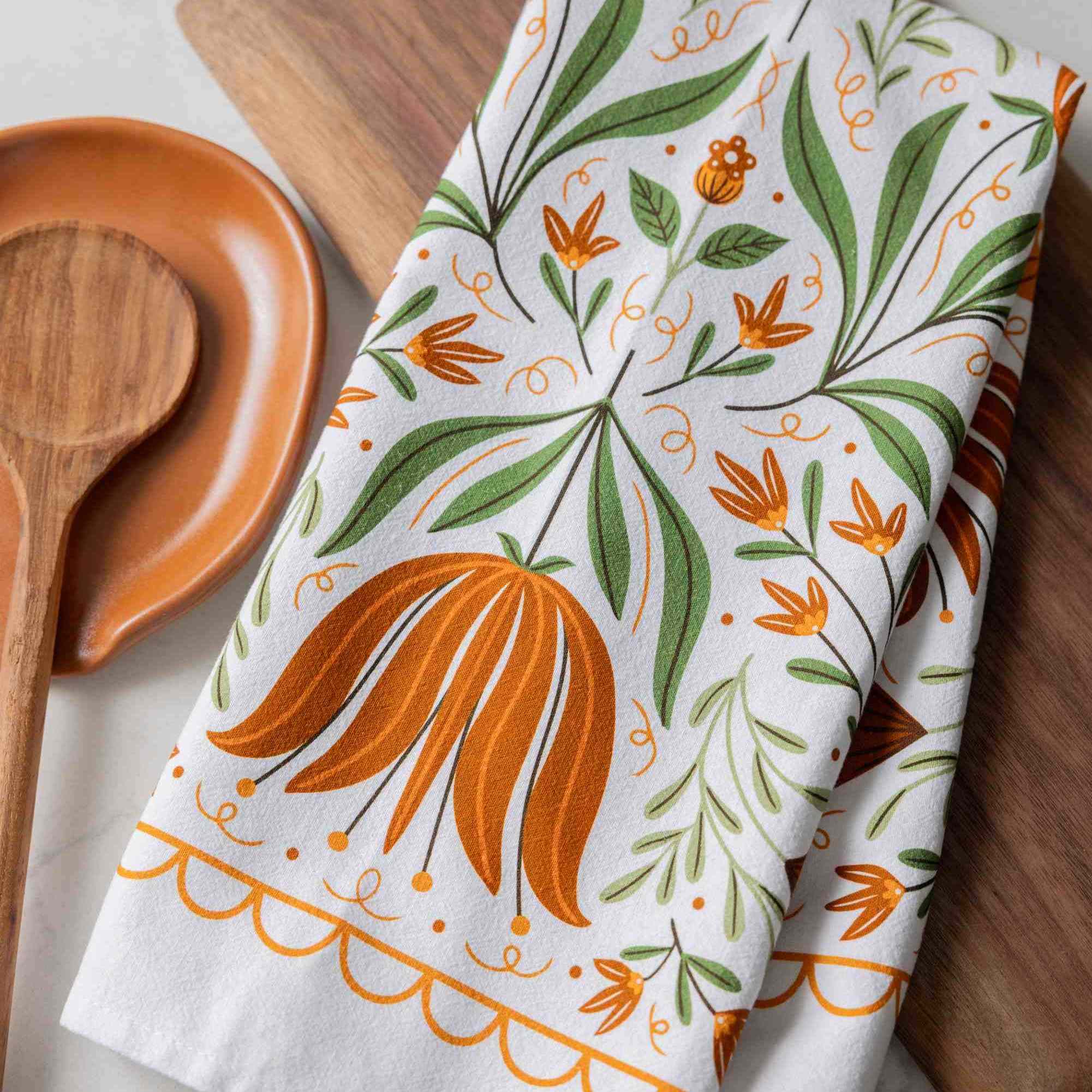 A white kitchen towel with a colorful botanical pattern of orange flowers and green leaves, styled on a wooden cutting board next to a wooden spoon.