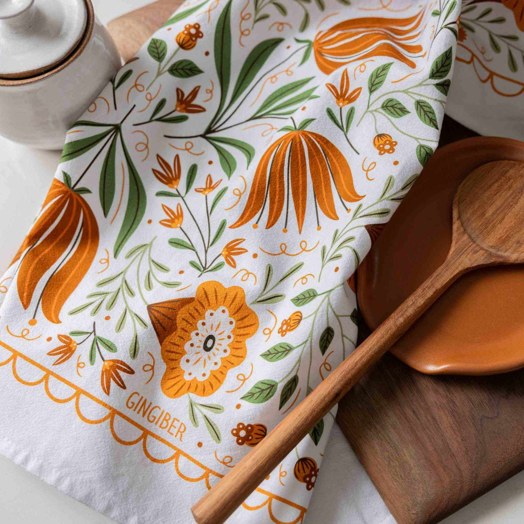 A white kitchen tea towel with a ginger-themed botanical pattern in orange and green, styled with a wooden spoon and a ceramic container.