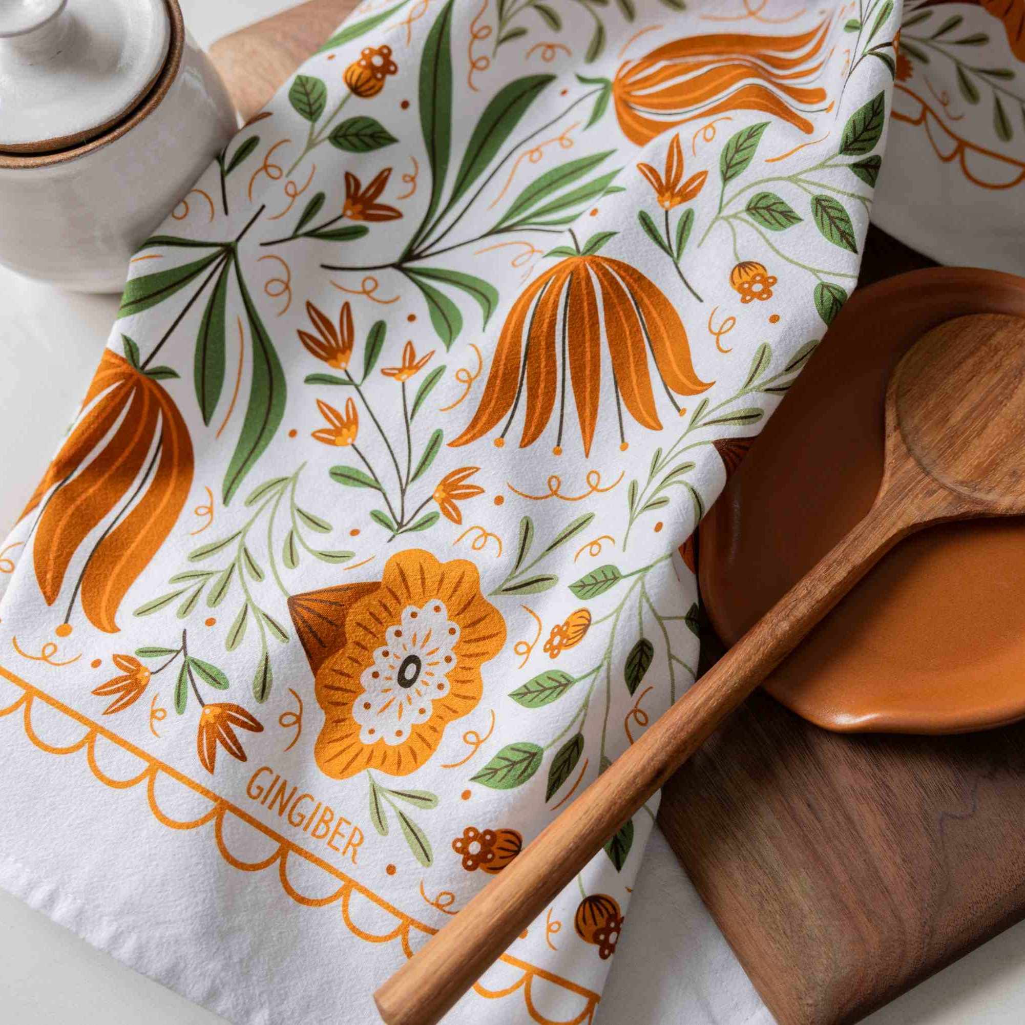 A white kitchen tea towel with a ginger-themed botanical pattern in orange and green, styled with a wooden spoon and a ceramic container.