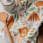 A white kitchen towel with a gingerbread and botanical pattern in orange and green, styled with a wooden spoon and a ceramic container on a wooden surface.