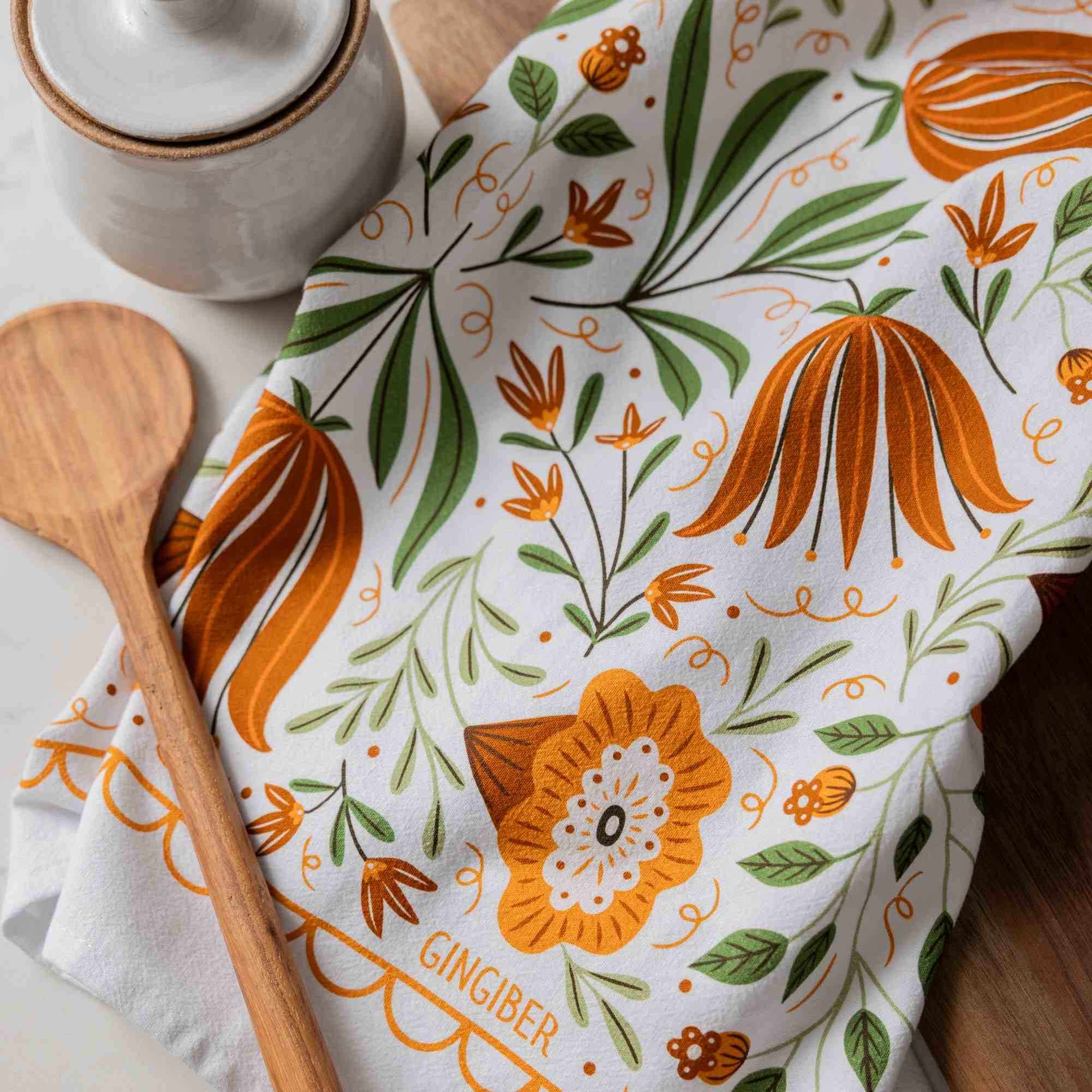 A white kitchen towel with a gingerbread and botanical pattern in orange and green, styled with a wooden spoon and a ceramic container on a wooden surface.