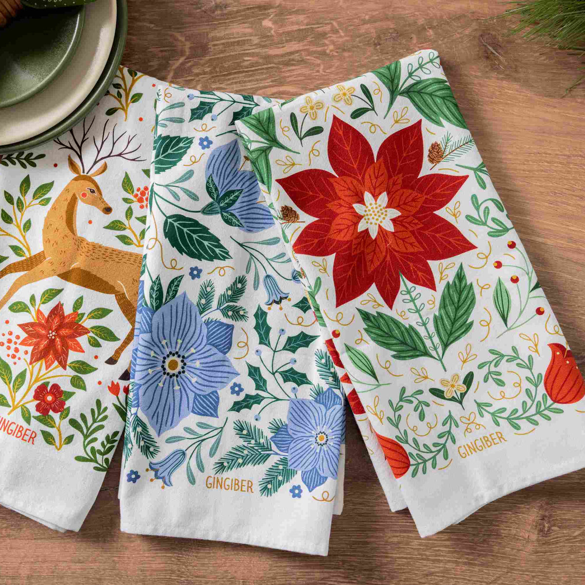 Three Christmas tea towels with festive printed designs, including a deer, blue flowers, and a red poinsettia arranged on a wooden tabletop next to a green napkin.