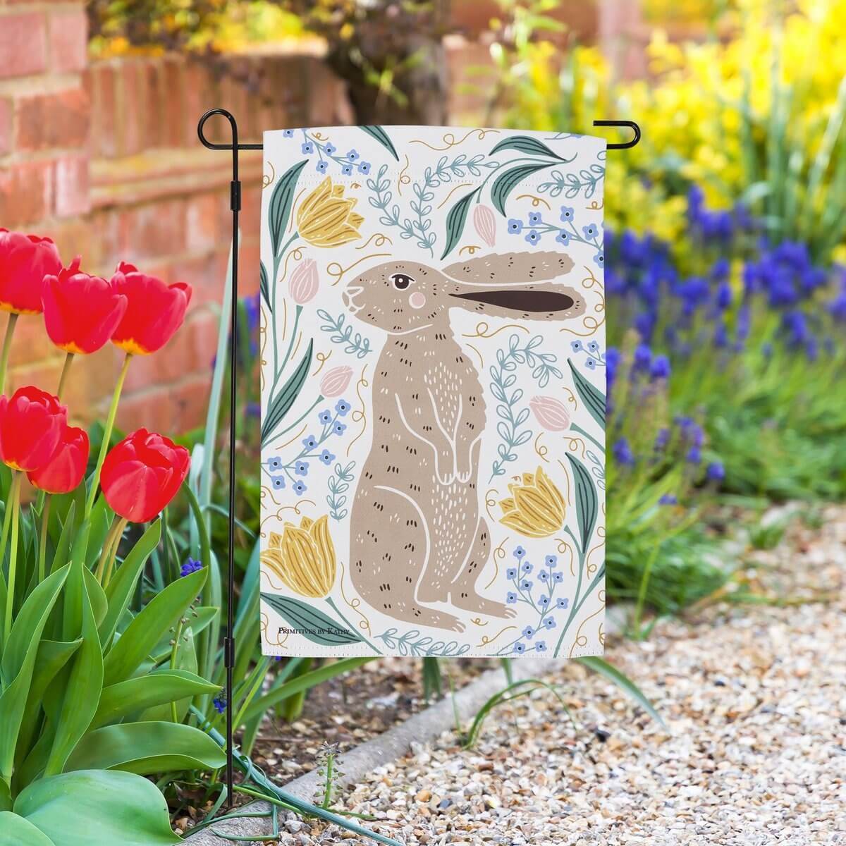 Decorative garden flag with a rabbit design in a garden setting with tulips and flowers.