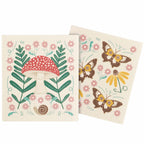 Two square cream-colored kitchen towels with whimsical nature illustrations, including a red and white mushroom, green leaves, pink flowers, yellow flowers, and brown butterflies.