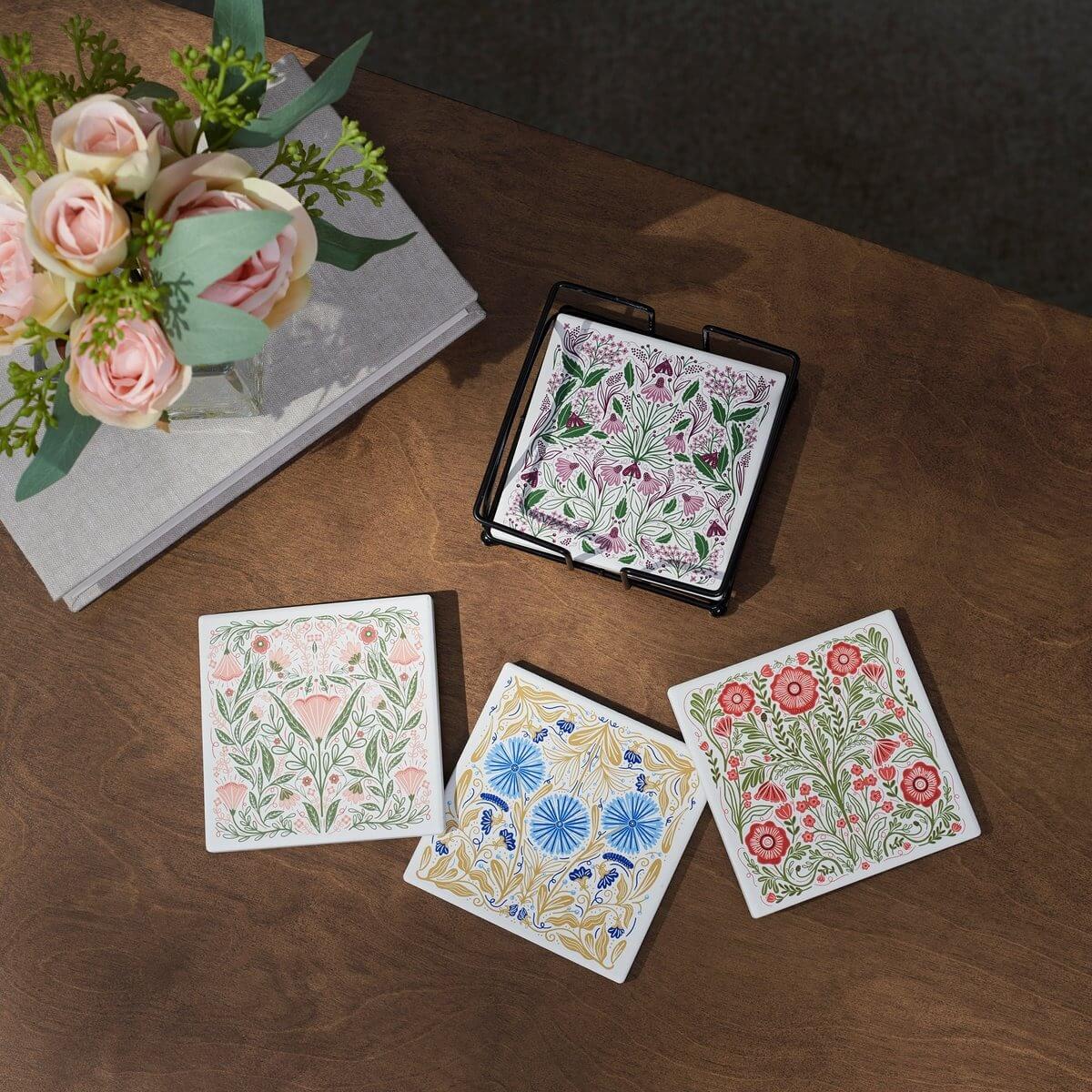 Set of floral coasters on a wooden surface with a book and flowers in the background.