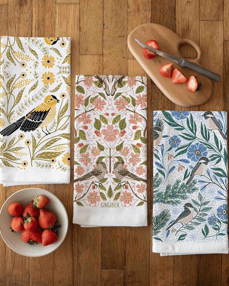 Three decorative kitchen towels with floral and nature-themed designs on a wooden table.