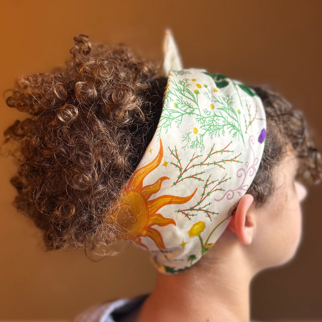 A close-up side view of a person wearing a white fabric headband with a colorful nature print featuring a yellow-orange sun, green leaves, and purple flowers. The background is a solid warm brown wall.