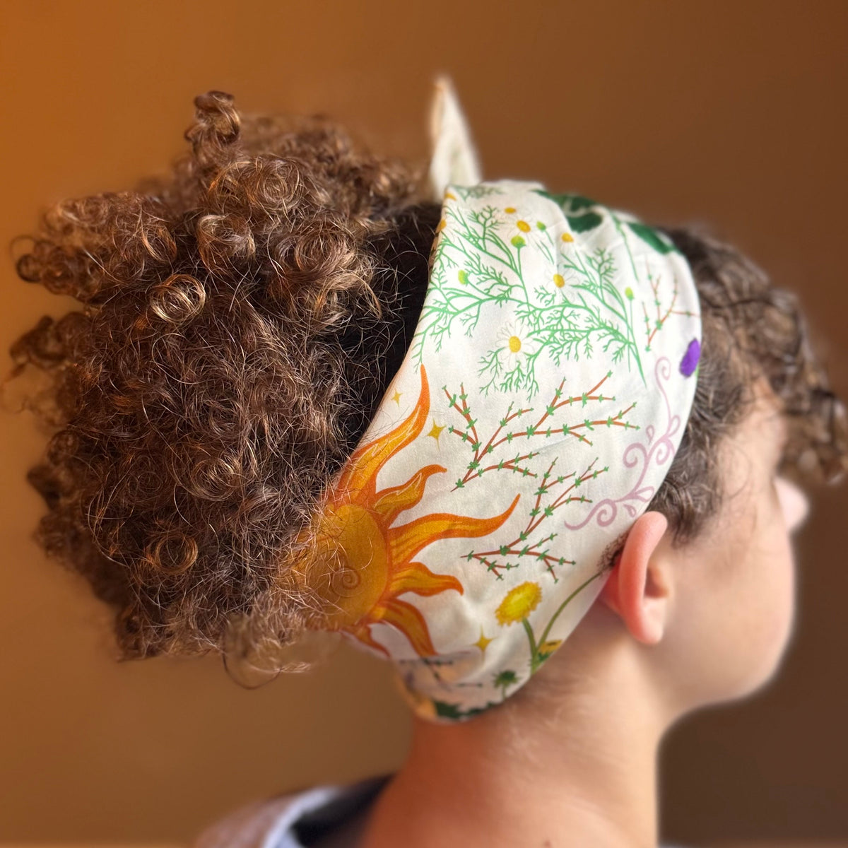 A close-up side view of a person wearing a white fabric headband with a colorful nature print featuring a yellow-orange sun, green leaves, and purple flowers. The background is a solid warm brown wall.
