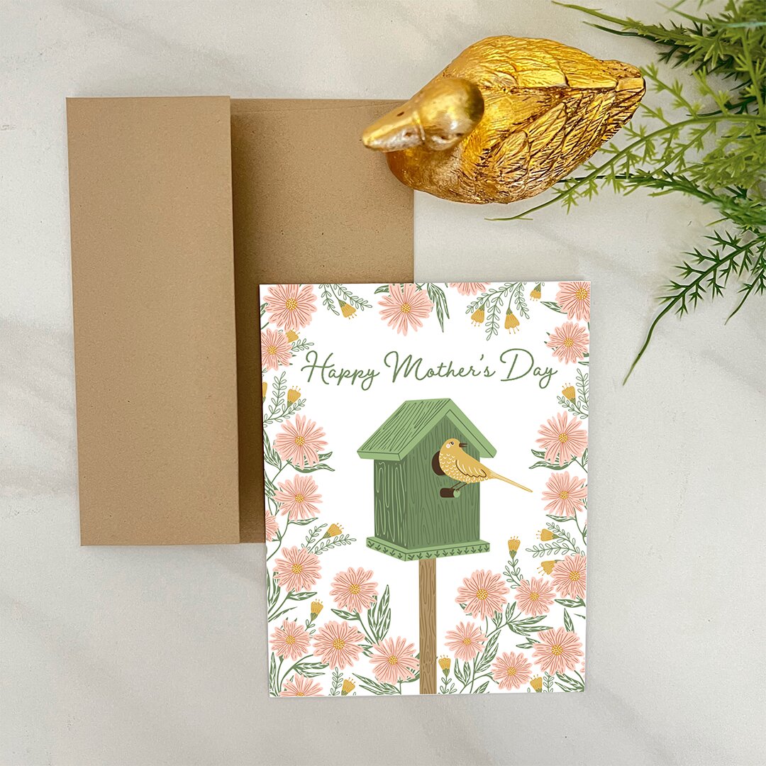 Greeting card with 'Happy Mother's Day' text and birdhouse illustration, accompanied by a gold mouse figurine and greenery.