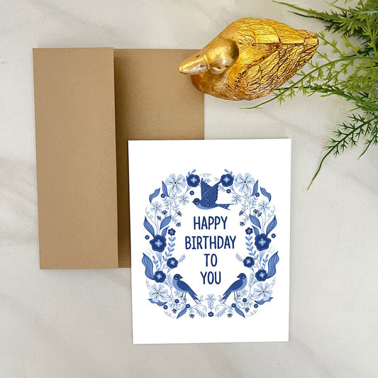 Birthday card with blue birds and flowers design, gold duck, and brown envelope on a light background