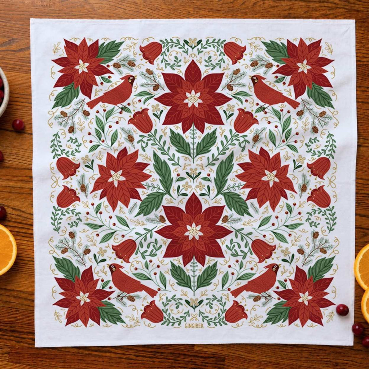 A square white kitchen towel or tea towel laid flat on a wooden table, featuring a festive Christmas pattern with red poinsettias, green leaves, and red birds. The towel is styled with holiday props including cranberries, orange slices, and a small bowl on a warm wood background.