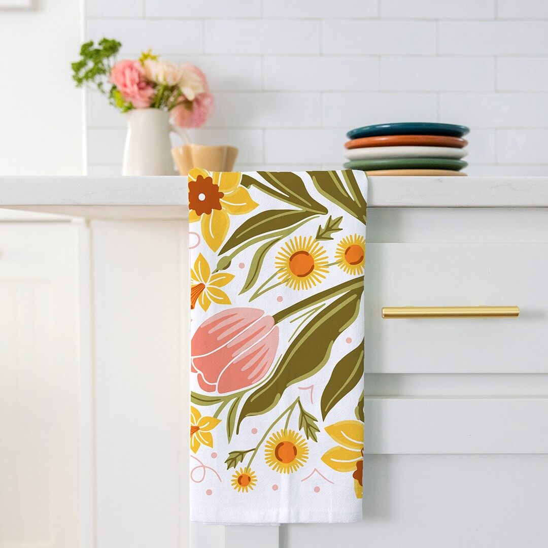 Floral towel hanging on a kitchen rack with a white tiled wall and decorative items in the background.