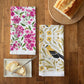 Two floral-themed kitchen towels on a wooden table with bread and butter.
