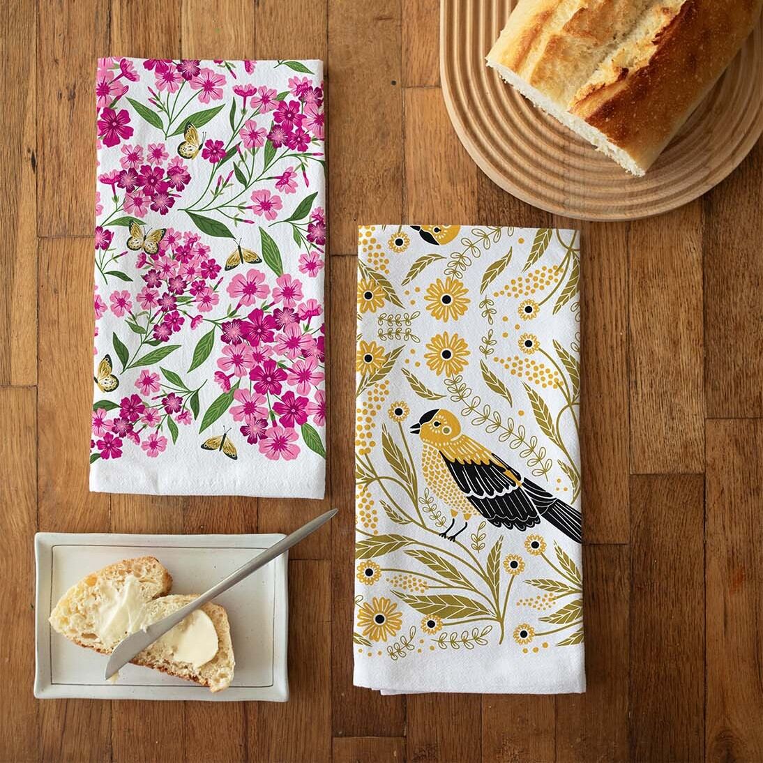 Two floral-themed kitchen towels on a wooden table with bread and butter.