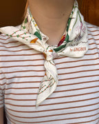A close-up of a person wearing a cream silk scarf with a ginger plant pattern and the text 'by GINGIBER' tied around their neck over a brown and white striped t-shirt.