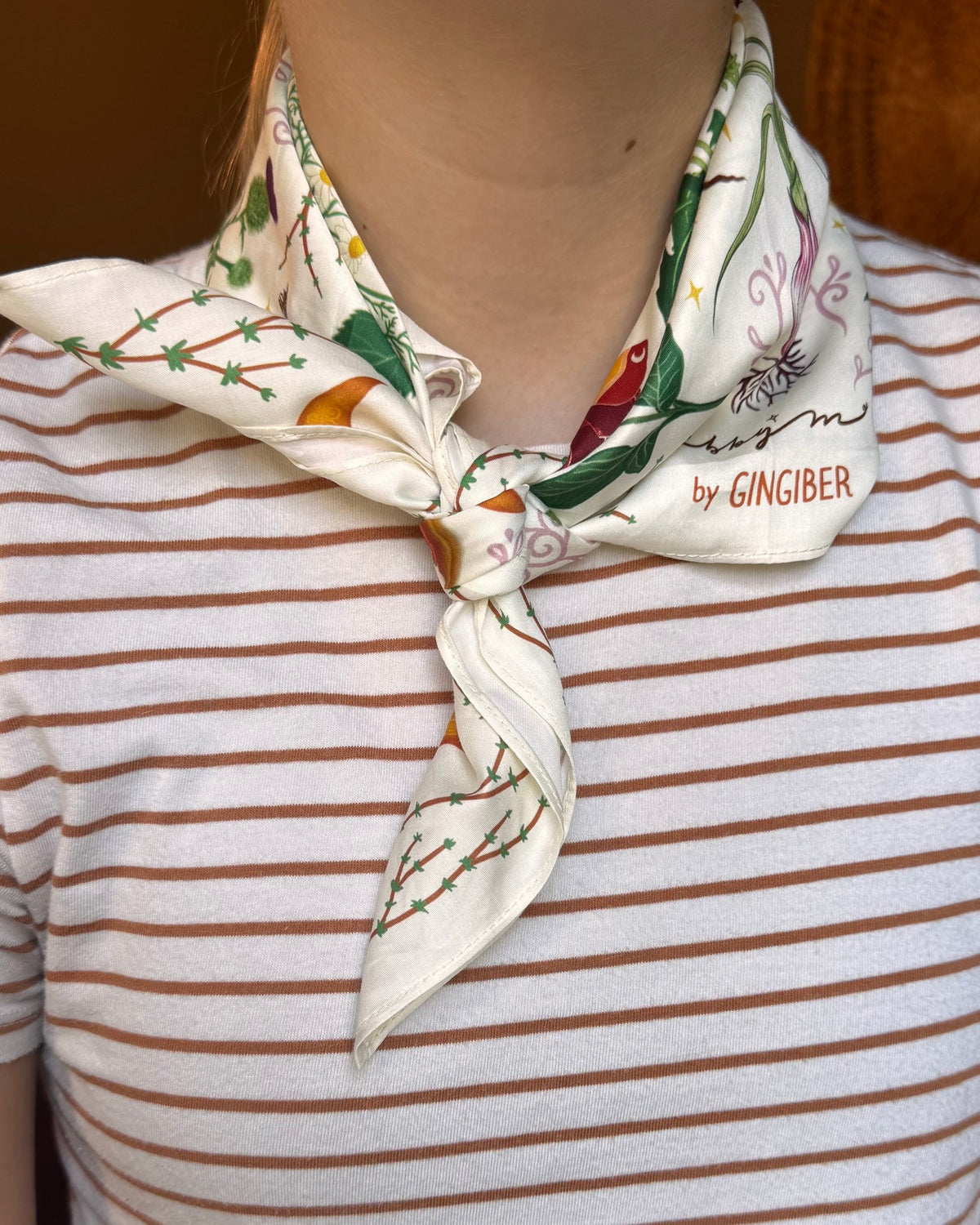 A close-up of a person wearing a cream silk scarf with a ginger plant pattern and the text 'by GINGIBER' tied around their neck over a brown and white striped t-shirt.