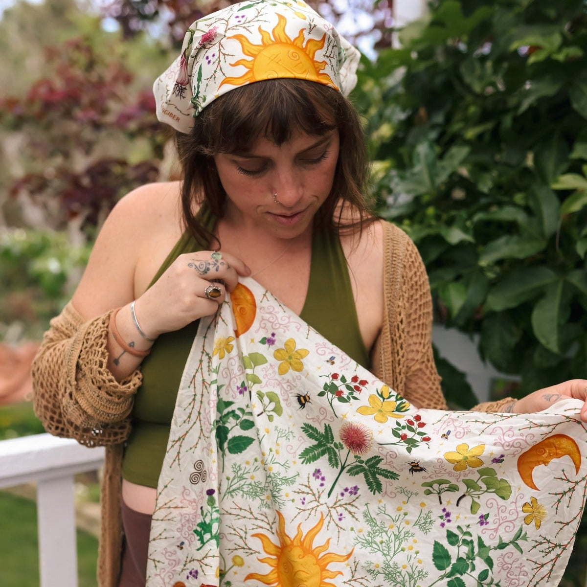 A woman outdoors holds up a square cream  bandana with a colorful embroidered sun, flowers, leaves, and crescent moons. She is wearing a green top, brown pants, and a beige cardigan, with a white railing and green foliage in the background.