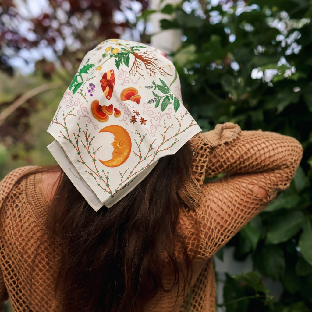 A woman viewed from behind wearing a cream-colored sun hat with colorful embroidered autumn motifs, including a crescent moon, acorns, and leaves, paired with a brown crochet knit sweater in an outdoor garden setting.
