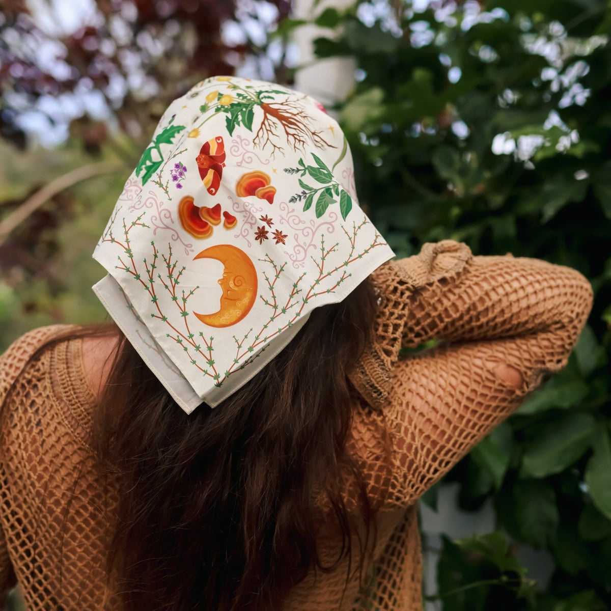 A woman viewed from behind wearing a cream-colored sun hat with colorful embroidered autumn motifs, including a crescent moon, acorns, and leaves, paired with a brown crochet knit sweater in an outdoor garden setting.