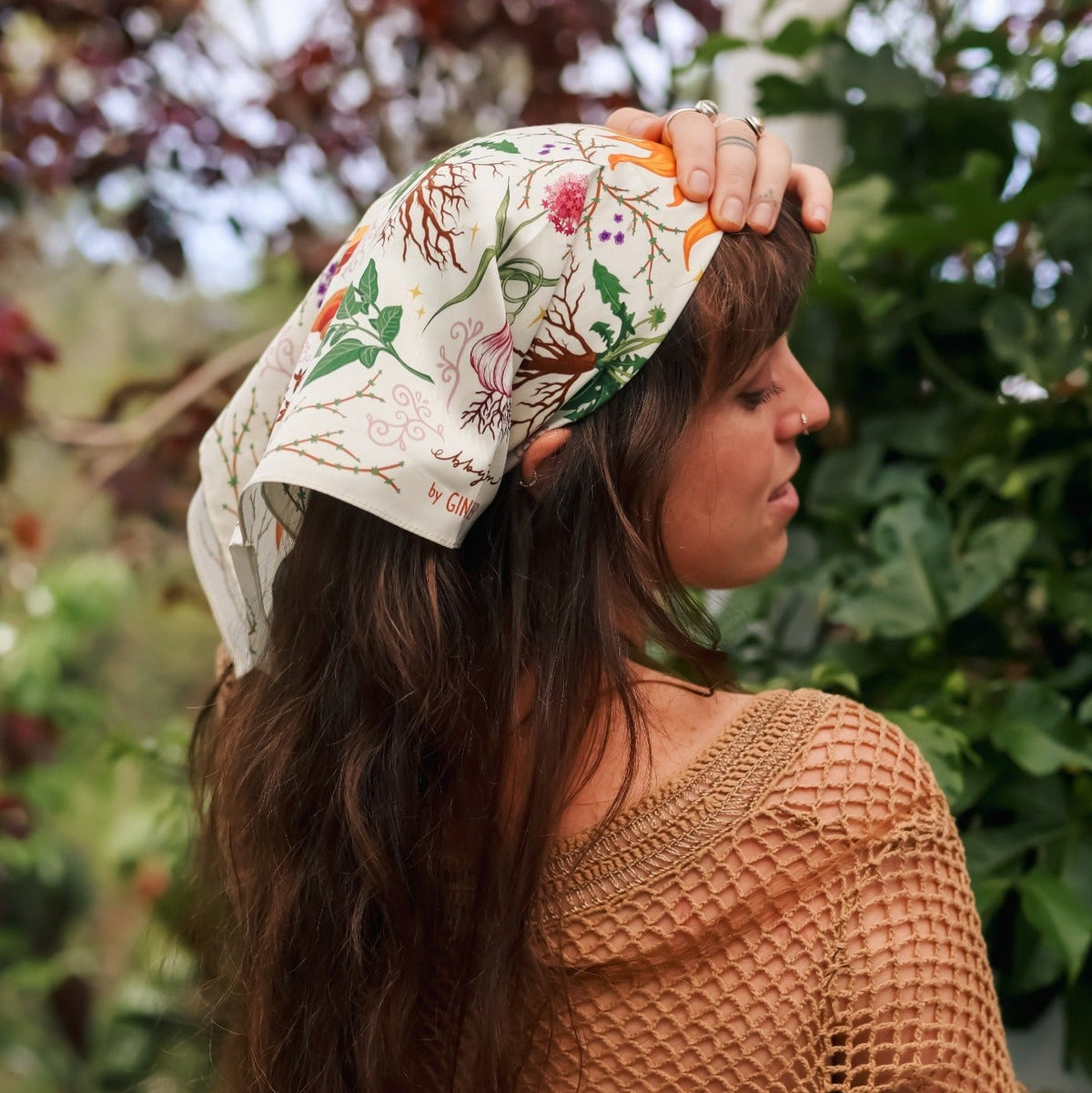 A woman with long brown hair is shown from the back and side, wearing a cream-colored headscarf with a colorful botanical pattern and a brown crochet knit top, set against a lush green garden background.