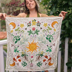 A woman outdoors holds up a square cream-colored textile featuring a colorful embroidered sun, flowers, leaves, birds, and crescent moons. The background shows a garden setting with green foliage and a white railing.