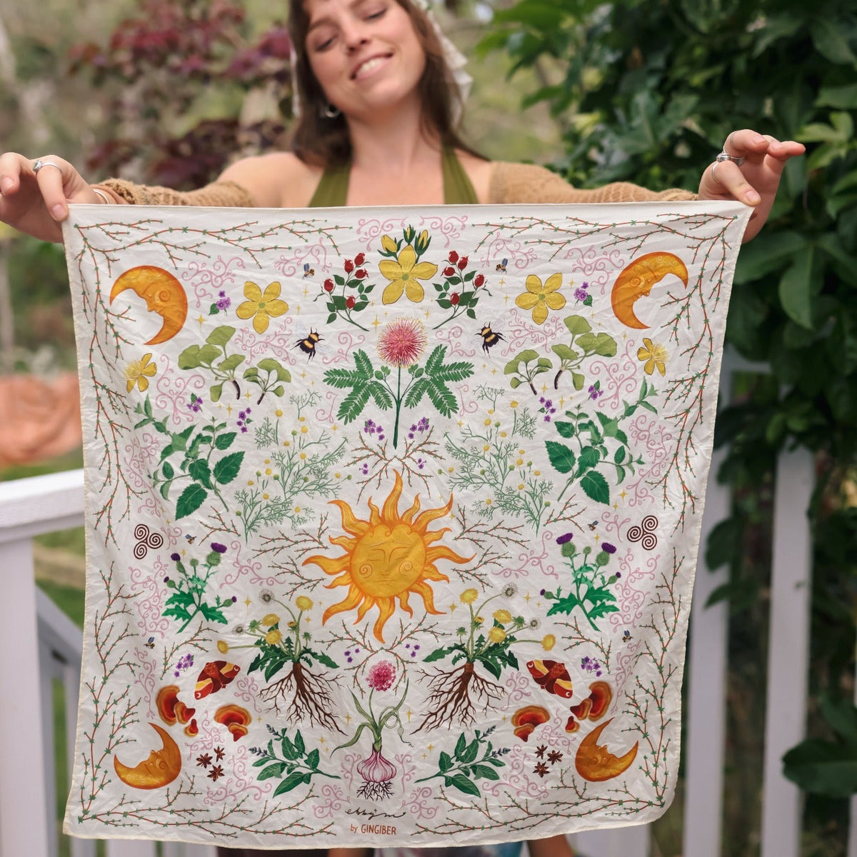 A woman outdoors holds up a square cream-colored textile featuring a colorful embroidered sun, flowers, leaves, birds, and crescent moons. The background shows a garden setting with green foliage and a white railing.